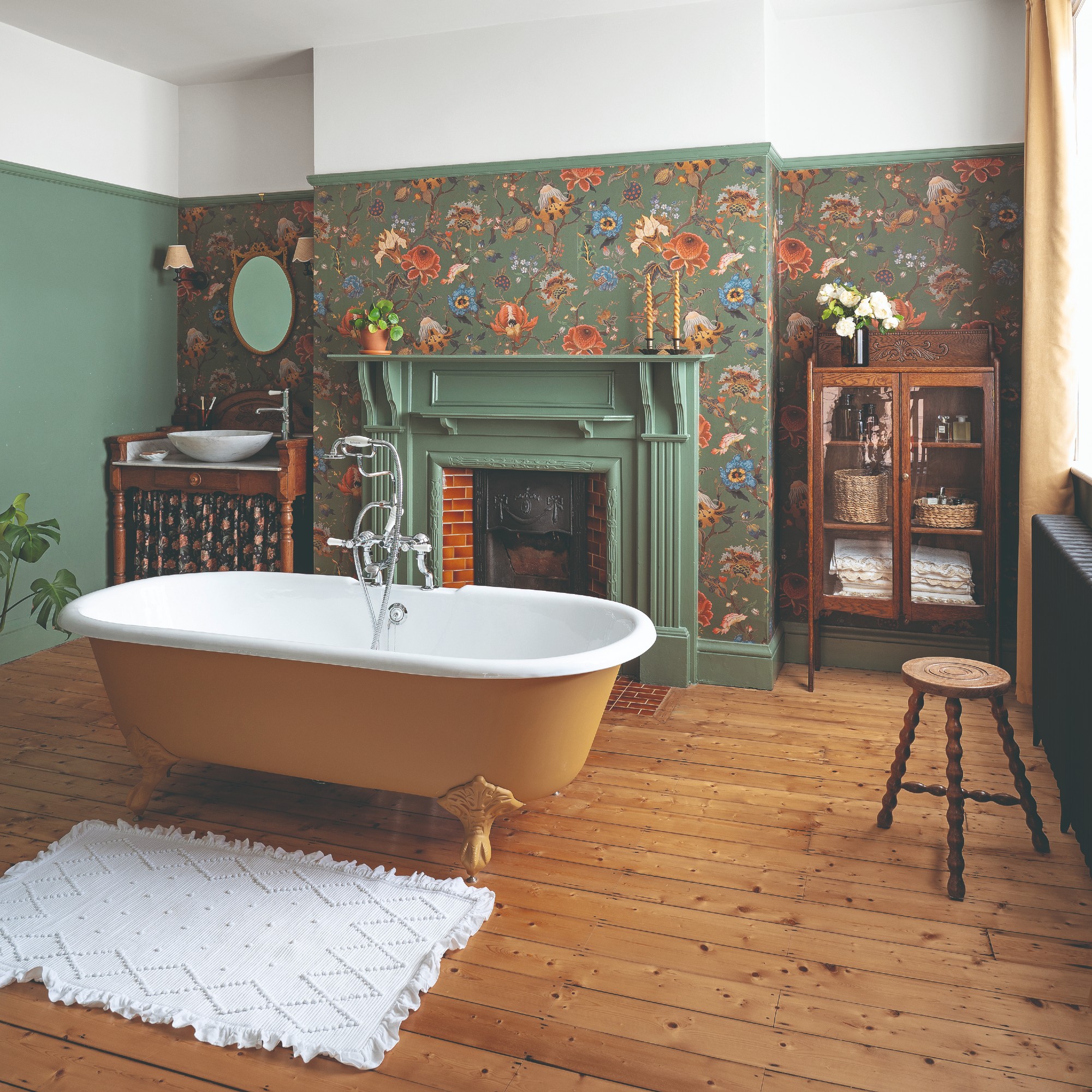 A bathroom with wooden floors painted in an earthy green and partly covered with a tonal floral wallpaper and a yellow bathtub in the middle of the room