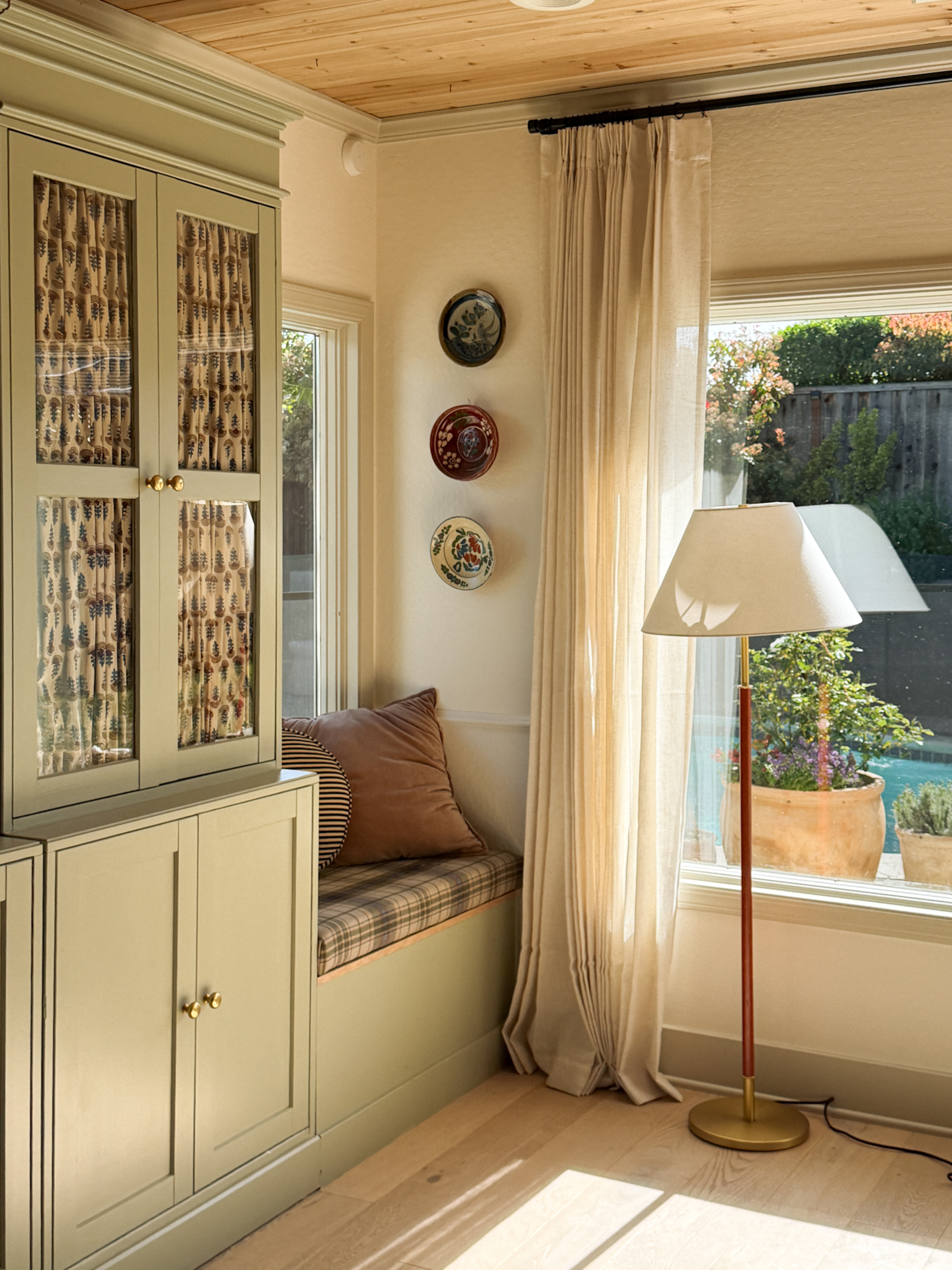 Image of the corner of a white room with a sage green built-in cabinet and sage green trim. There is a bench seat with plaid cushions, plates hanging on the wall, and a floor lamp.