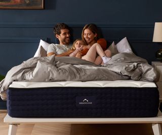 A man, a woman, and a child, lying on their backs on a DreamCloud mattress against a navy wall.