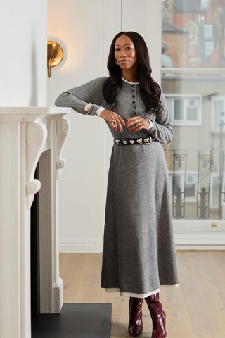 A woman standing inside her home wearing a gray cardigan over a white T-shirt, a matching gray knit skirt, a black studded belt, red knee-high boots, and silver cuff bracelets.