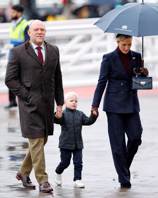 Mike Tindall, wearing a brown coat and red tie, and Zara Tindall, wearing a blue pinstriped suit and holding an umbrella, holding hands with Lucas tindall, who is standing in between his parents and scowling