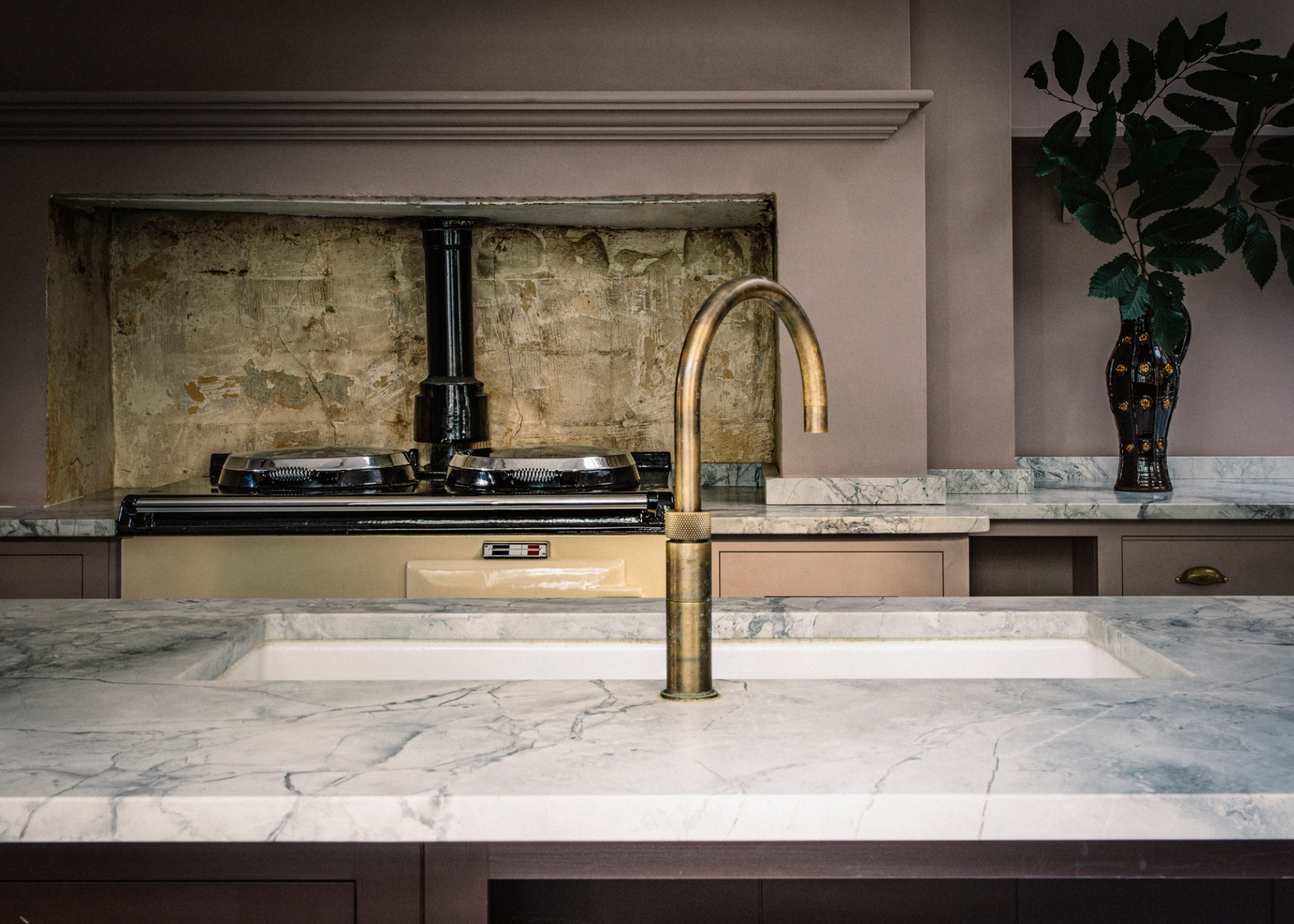 A close up of a brass kitchen tap with a natural patina, postioned over a white and grey marble countertop with brass splashback for the cooker in the background