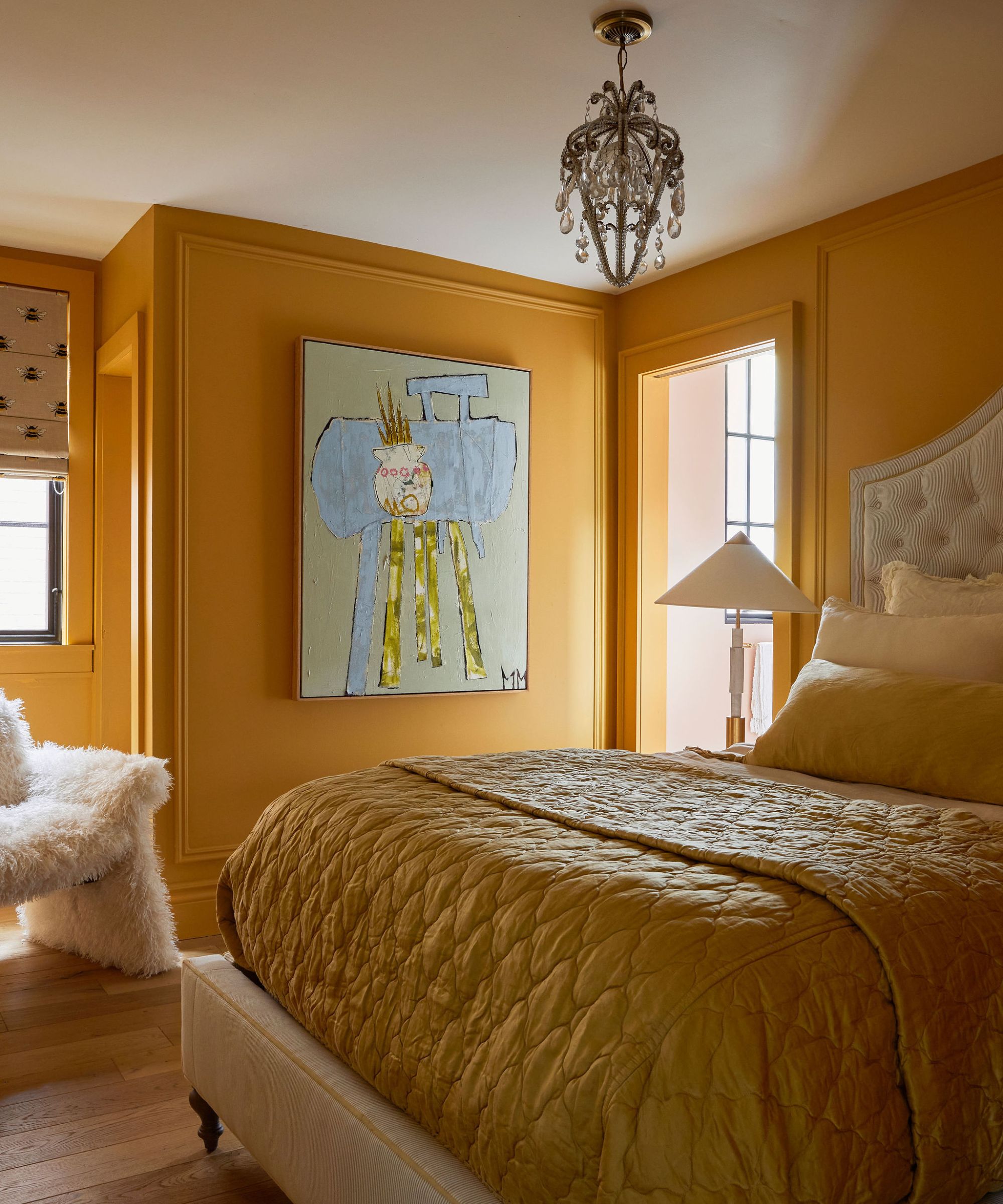 Yellow bedroom with a large bed, also covered in yellow bedding. A piece of playful artwork is on one wall and a furry chair sits in the corner under a window
