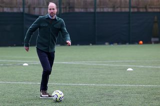 Prince William wearing a green shacket and blue pants kicking a soccer ball along grass and running