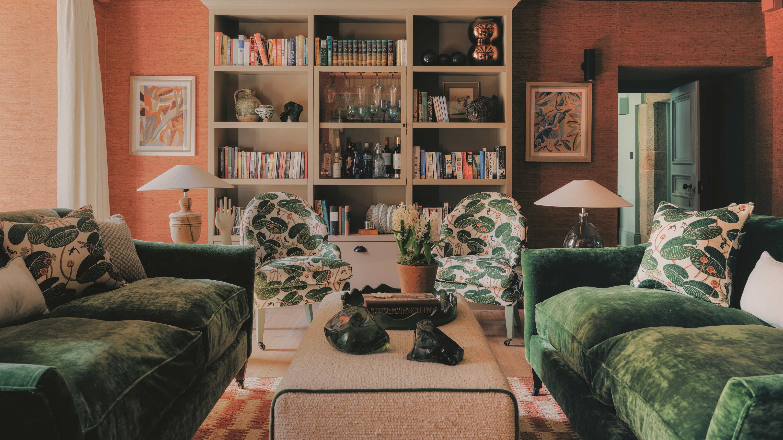 a soft orange living room with green velvet couches, a beige painted bookcase and palm leaf printed armchairs