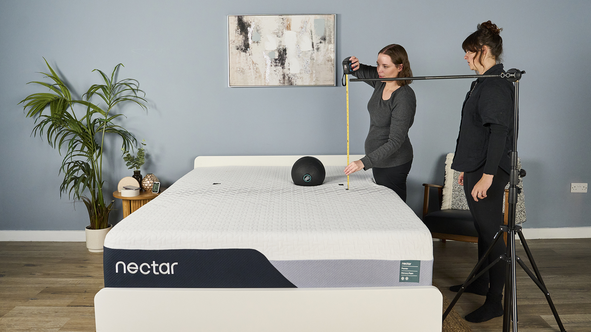 In this image mattress review writers Ruth Jones and Lauren Jeffries prepare to perform a motion isolation test using a slam ball on a memory foam mattress. They stand on the left hand side of the mattress and use a tape measure to position a rig at 24 inches, the height from which they'll drop the ball. A 5kg slam ball is currently resting on the mattress, to help them determine the drop zone