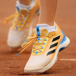 Someone wearing Adidas trainers on a sand tennis court