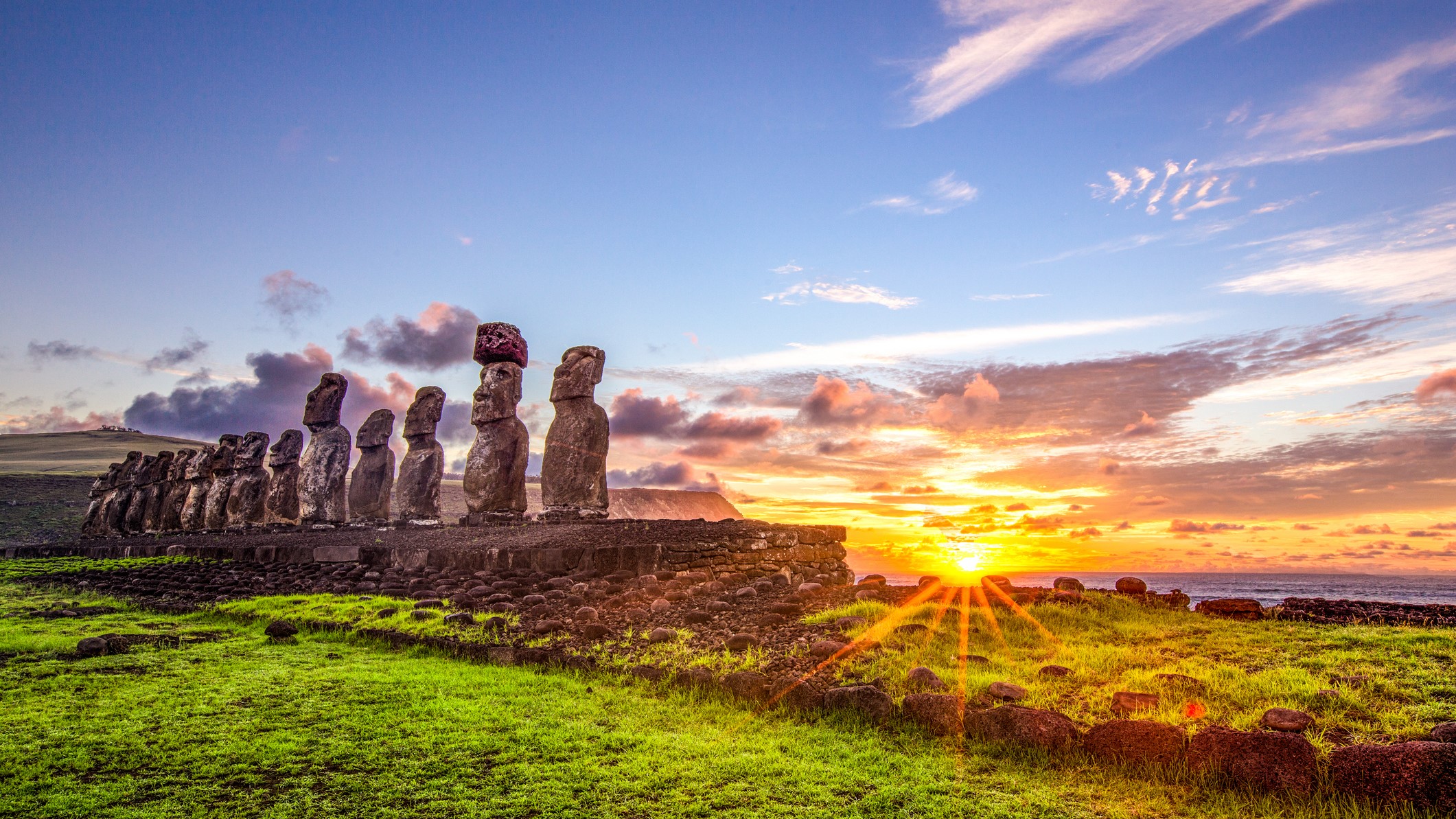 a row of moai statue heads are in the foreground with a rising sun over the ocean in the background.