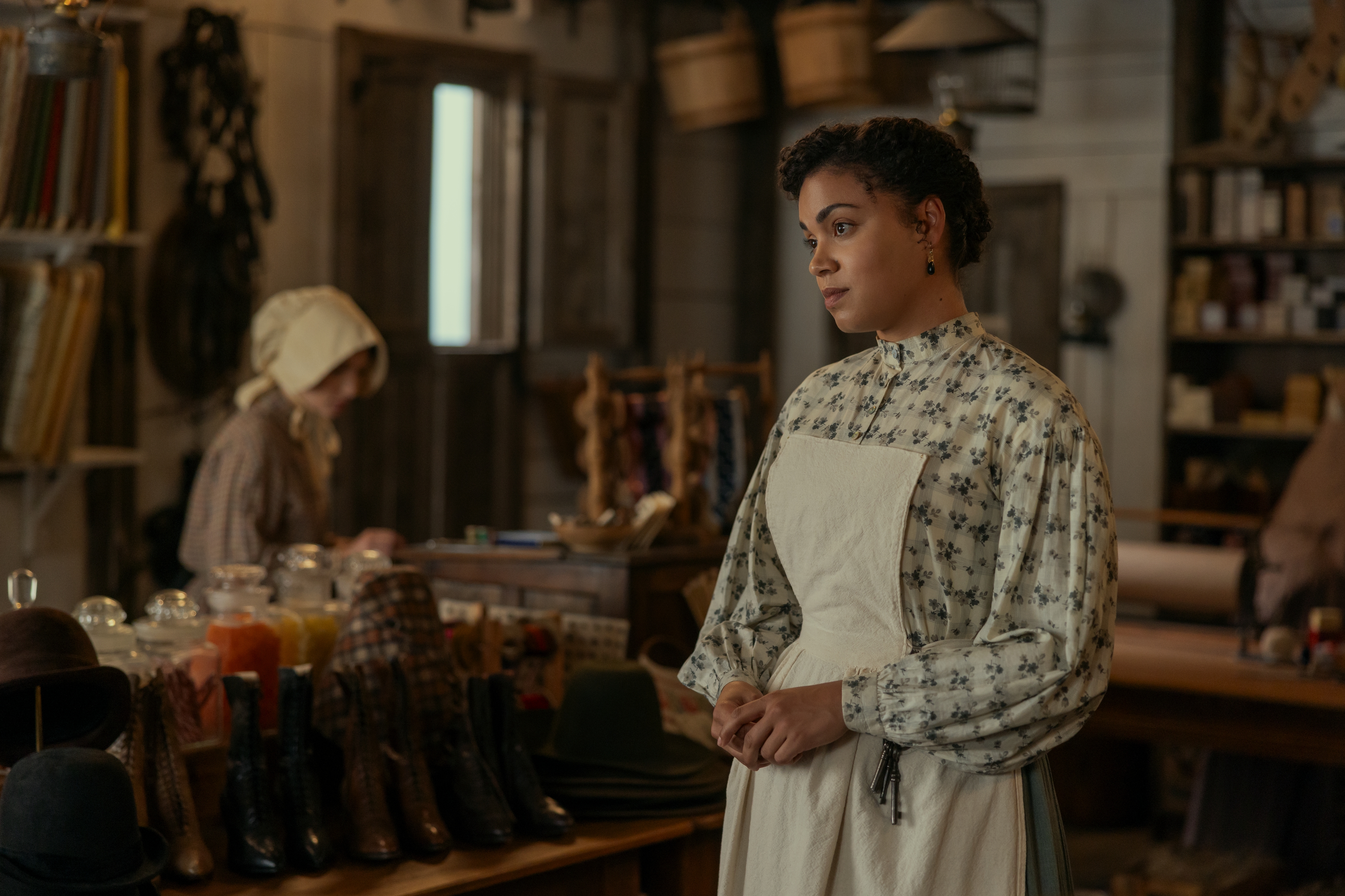 Actress Barrett Doss as Emily Henderson wears an apron while standing in a 19th-century general store, as a girl with a bonnet shops behind her, in Netflix's 'Little House on the Prairie.'