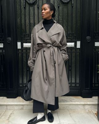 British influencer Marilyn Nwawulor-Kazemaks poses in from of a black door in London wearing a black turtleneck sweater, gray belted trench coat, cropped black pants, white socks, and black ruched loafers