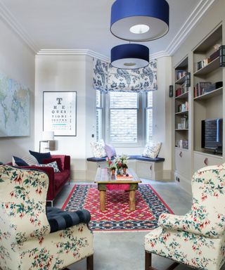 A bright living room with clashing colors and patterns, with a rug, two matching floral cream armchairs, a burgundy velvet sofa, color drenched open shelving filled with books, a wooden coffee table and large bay window with blue cushioning to match the two blue lampshades hanging from the ceiling