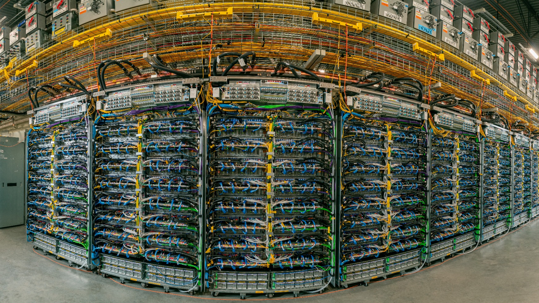 Rows of Google Ironwood TPU racks in a data center, with blue, green and yellow cabling under orange overhead cable trays.