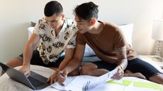 A couple look at paperwork and a laptop together while sitting on a bed.