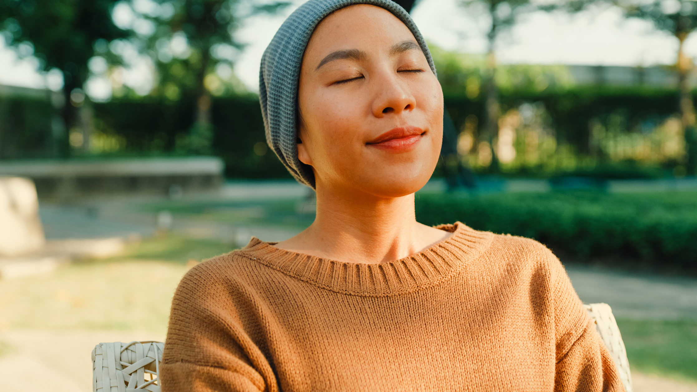 A woman sits in the sunshine in a garden. She has her eyes closed and wears a peaceful expression on her face