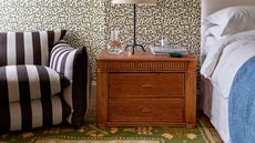 Lucy Williams Home x Pelican House rug collaboration showing a green floral flatweave rug in a floral wallpapered bedroom with white linens, a vintage bamboo nightstand and a black and white striped chair