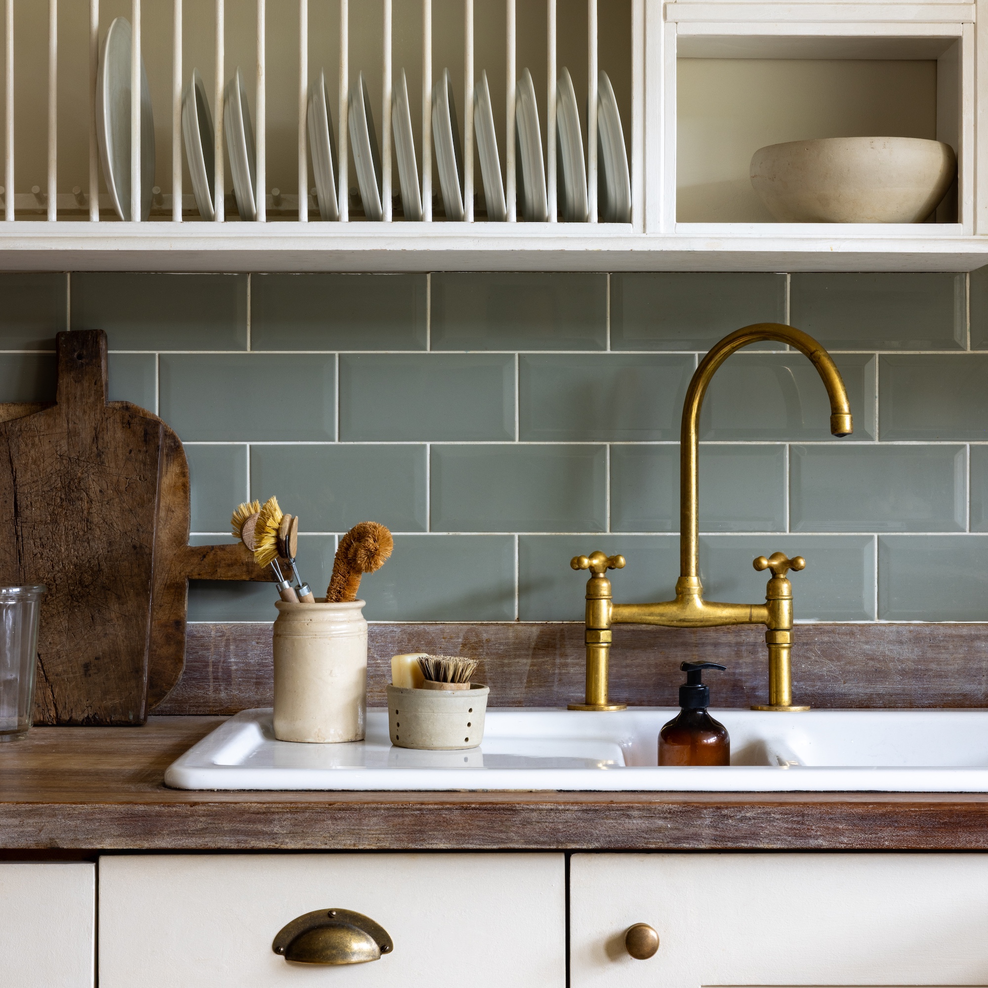 white ceramic inset sink with brass mixer tap and sage green metro tiles behind and white plate wallmounted plate rack above