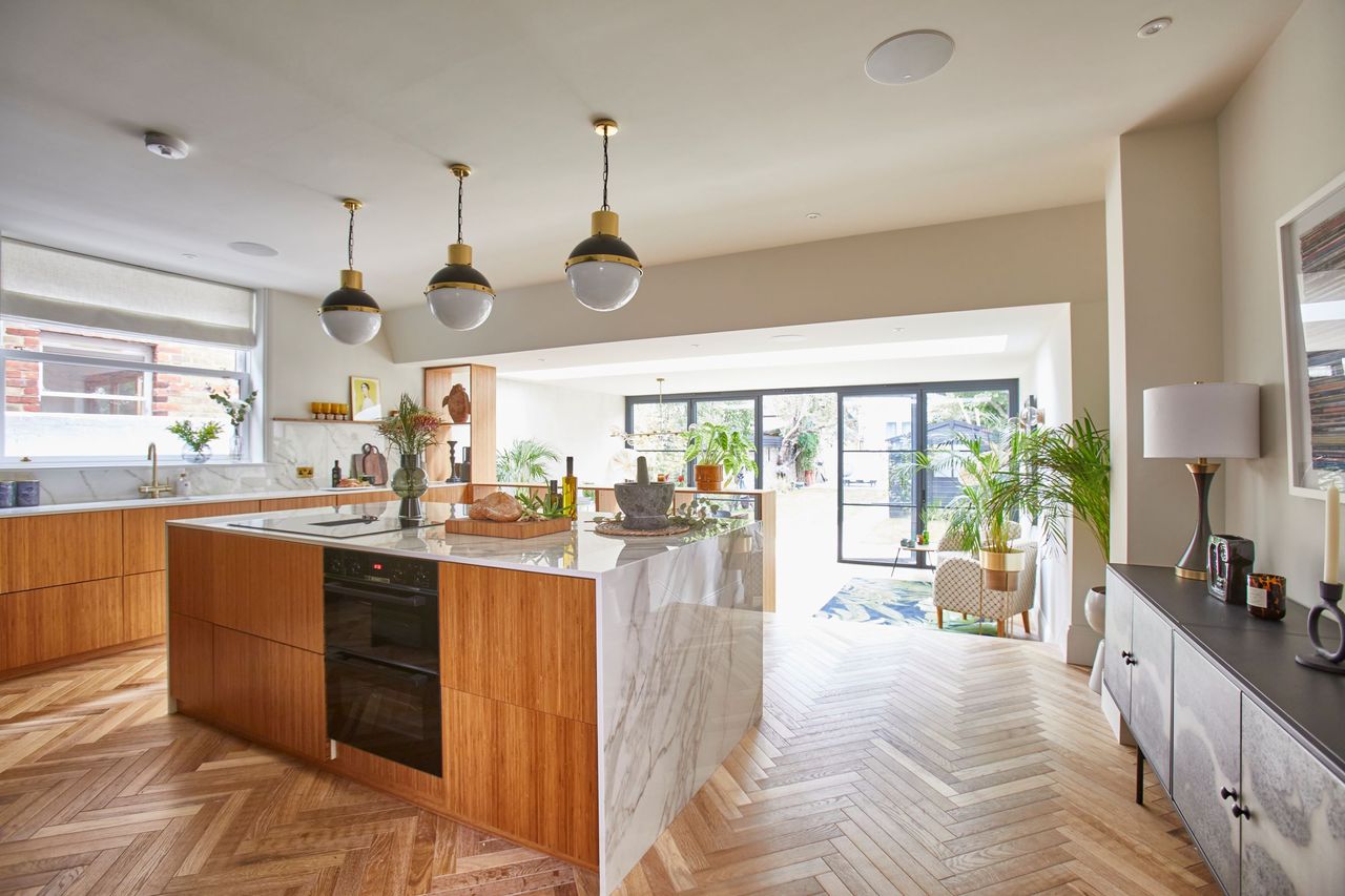 The kitchen-diner has wooden flooring and a kitchen island with an induction hub with three ceiling lights above with white marble worktops in the kitchen