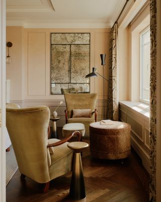 Backed by an antique glass mirror, two velvet upholstered wingback chairs sit in front of a window.