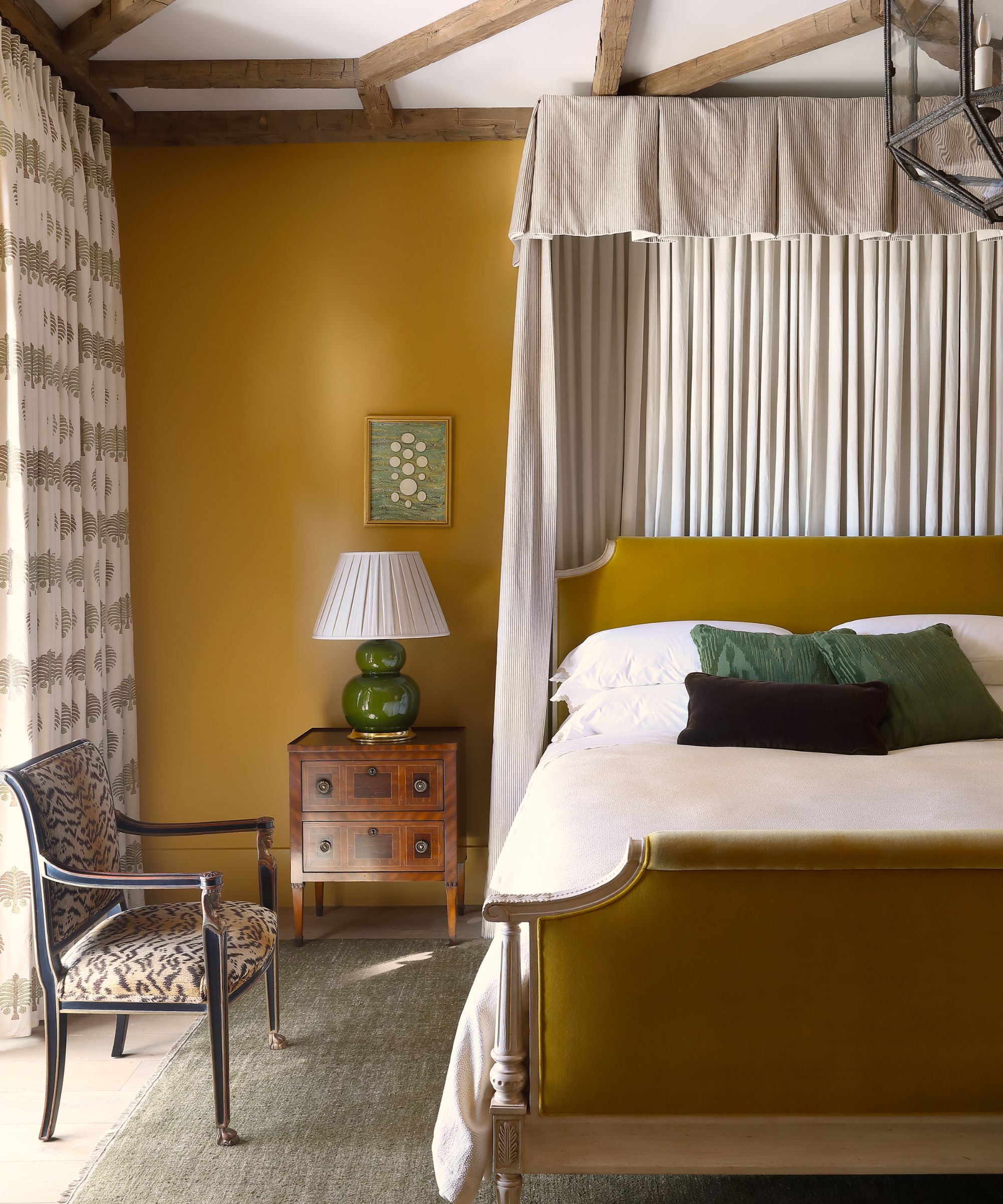 Bedroom with beamed ceiling, mustard velvet bed frame, mustard walls, antique bedside table and chair, and bed canopy
