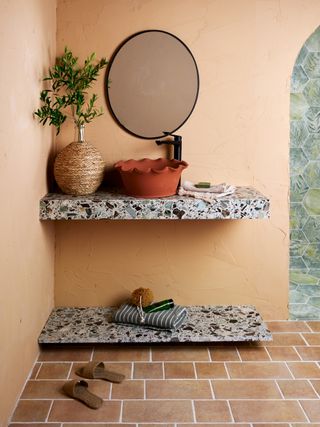 Image of a light peach-painted bathroom with dark and light blue terrazzo tile on both the vanity top and the floating shelf underneath. There is a red clay sink installed on the vanity, and a round mirror hanging over the sink.