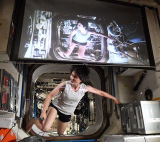 astronaut samantha cristoforetti floats in a space station module in a white tank top, hanging on to a box on the right. above her is a tv screen with sandra bullock in the same position in the movie gravity