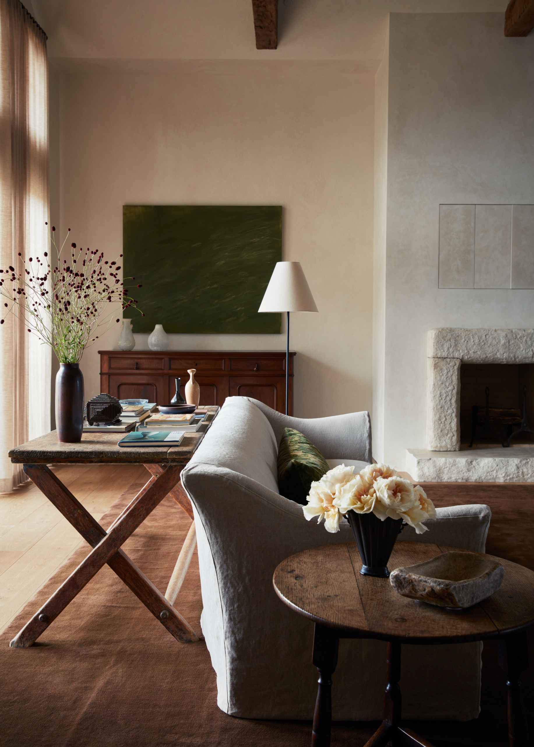 Folding wooden table behind a beige-covered sofa topped with books, vases and flowers, with a dark wood sideboard and green painting above in the background