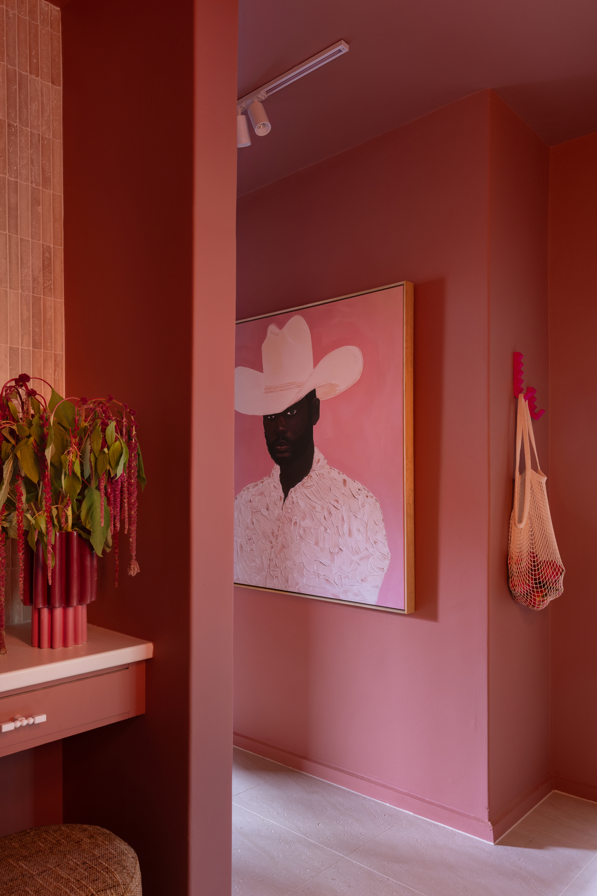 Image of a hallway drenched in pink with a painting of a cowboy wearing a white cowboy hat. There is a hot pink hook on the wall holding a next bag.