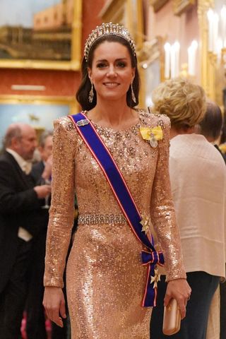 Kate Middleton smiling and wearing a pink sequined gown and blue sash along with a diamond tiara