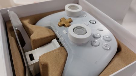 A GameSir Cyclone 2 controller in the phantom white colourway wrests in the cardboard insert inside of its box. This is the Charging Station edition, and the aforementioned cradle can also be seen in the box. The cradle's golden pins that attach to the underside of the gamepad for charging are just about visible.