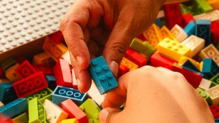 An open box of LEGO® Braille Bricks. A hand holds up a blue LEGO Braille Brick. Another hand is poised to pick a LEGO Braille Brick from the box.