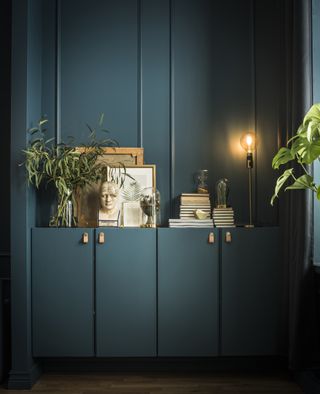 dark and stylish Ikea cabinets painted teal/blue with small handles by ikea, with dark paneled walls behind and frames, books, plants, and an exposed lightbulb lamp on top of the cabinet