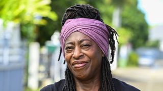 A close-up of Dorna Bray (Judith Jacob) in the street outside her home. Her hair is in dreadlocks piled high on her head, with a purple scarf wrapped around her head.