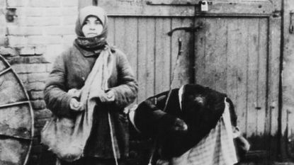 black and white photo of two women, one with satchel, bundled up the other in a jacket bending to the ground