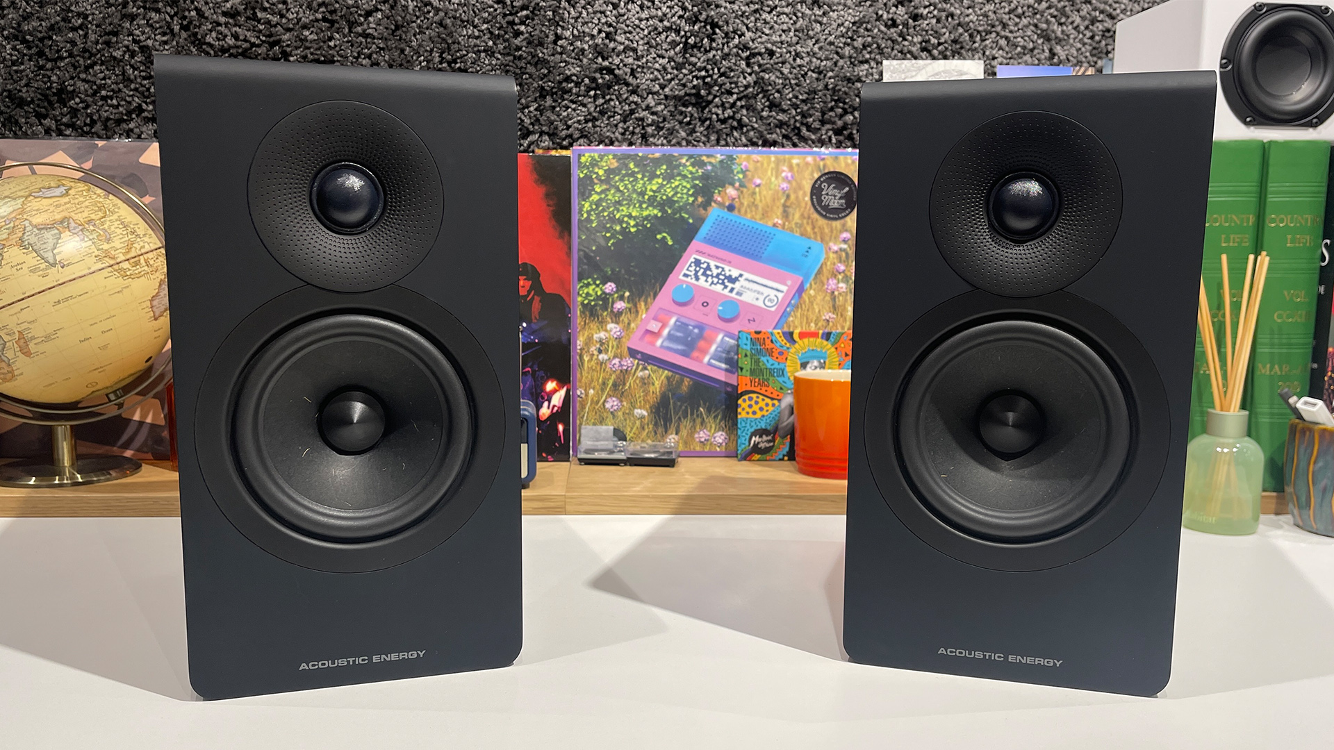 Acoustic Energy AE300 Mk2 standmount speakers on white surface in front of busy bookshelf