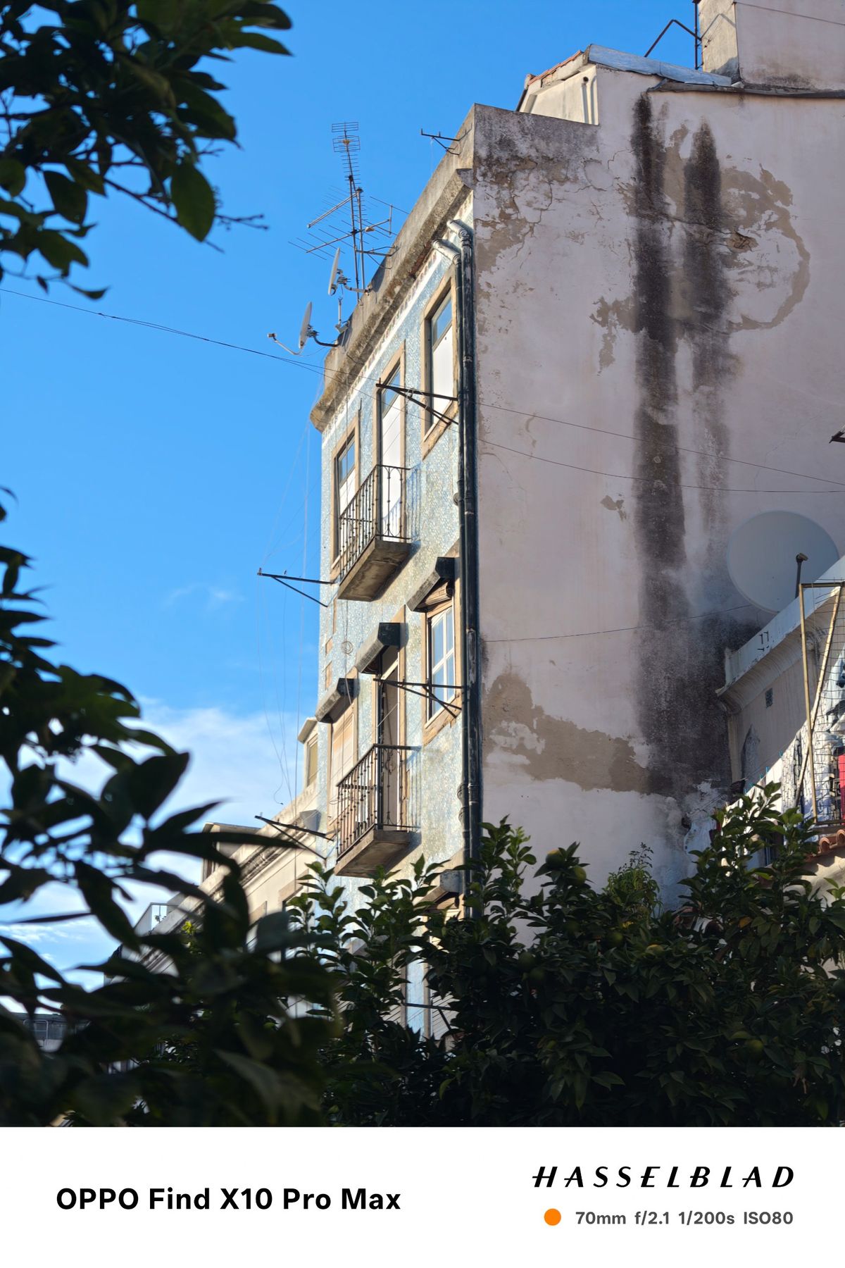 Weathered Lisbon apartment building with balconies, antennas and satellite dishes framed by trees.