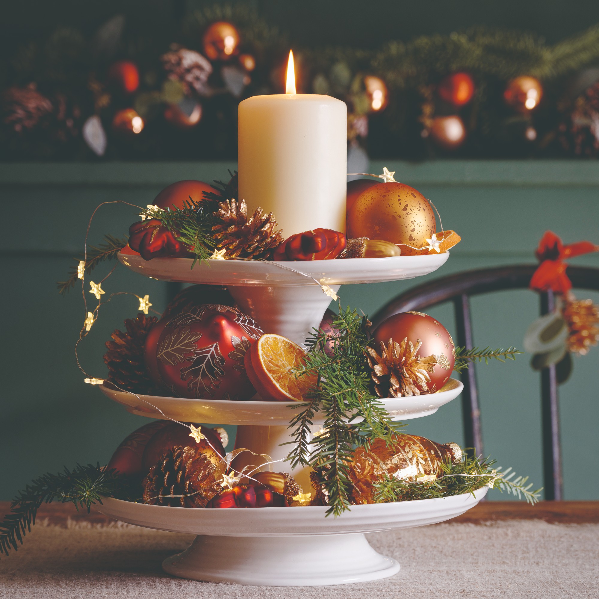 A dining table decorated for Christmas with a centrepiece made with a 3-tier serving platter displaying baubles, pinecones, fairy lights and a pillar candle on top