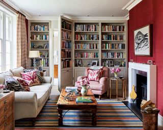 Living room with floor to ceiling gray shelving units, filled with books, large cream sofa and pink armchair, large striped multicolor rug, wooden coffee table. modern fireplace with magenta wallpaper on chimney breast