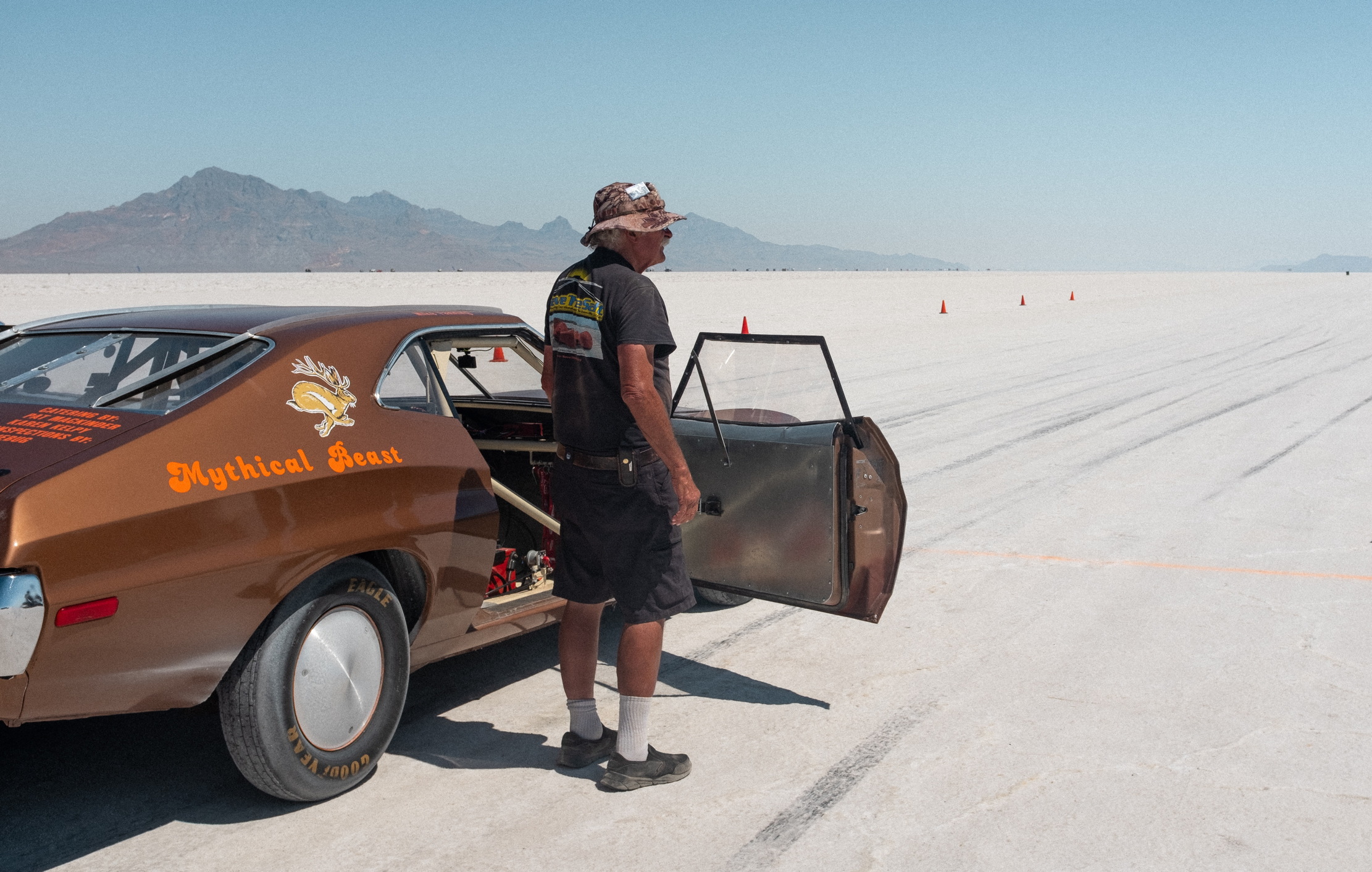 A man stands next to a bronze Oldsmobile, wistfully surveying the salt flats