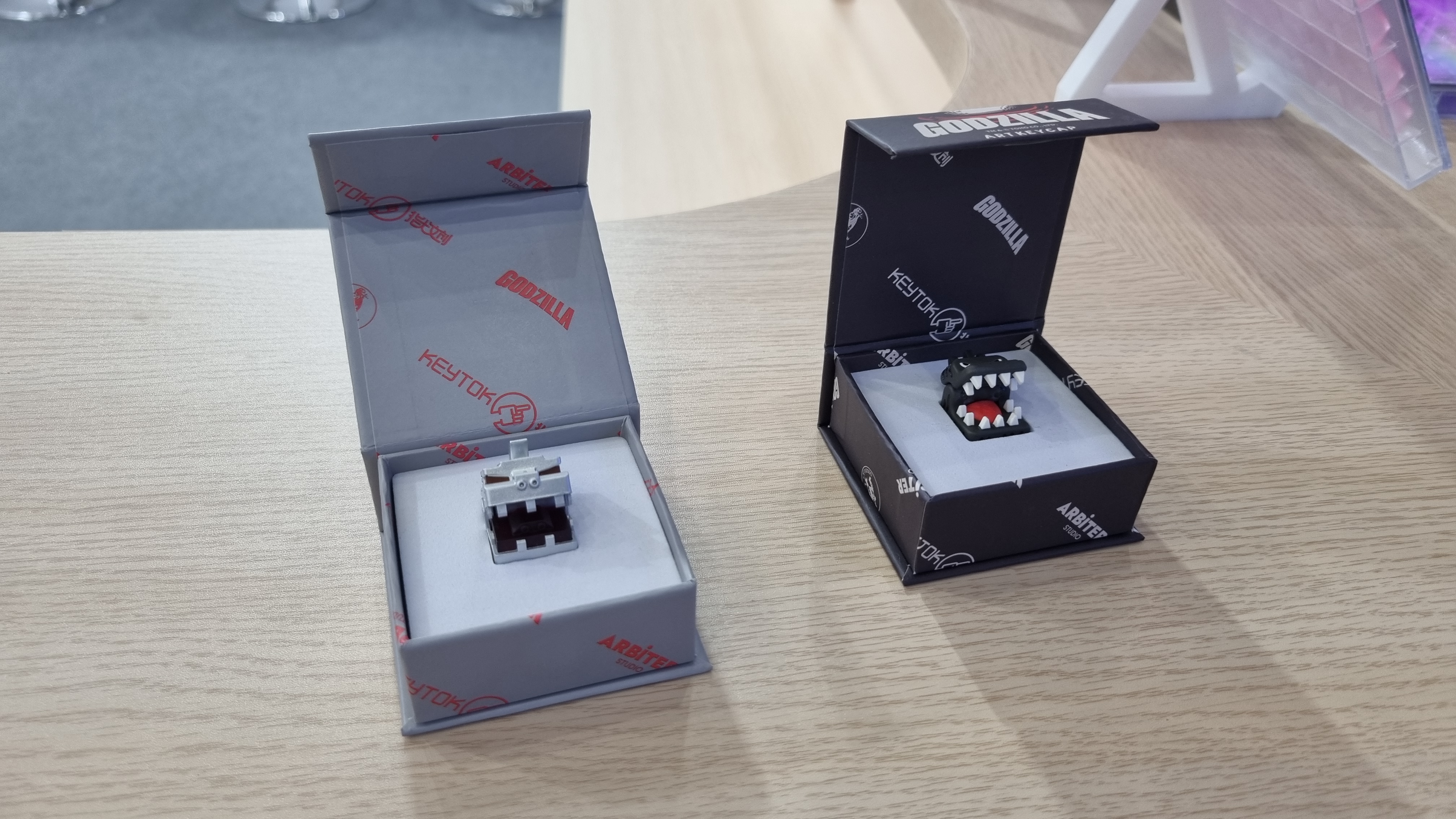 Two Godzilla keycaps in boxes on a table
