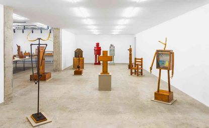 Gallery space, white walls and ceiling with lights, concrete floor and archway, floor standing wooden art pieces placed around the floor, additional room in view with display tables of wooden art pieces 