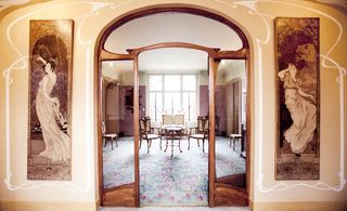 Interior room inside Perrier-Jouët house, wooden and glass arch doorway, long portrait of females on either side, floral carpet through the arch, high white ceiling, pink walls, small round table and chairs, large window letting in daylight at the far end