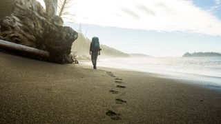 Person walking along beach with backpack, leaving a trail of footprints