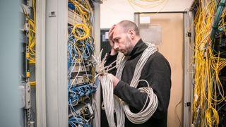 A sad technician wrapped in server cables assesses a server in a data centre with dismay