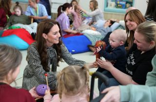 Kate Middleton wearing a black plaid dress sitting on the floor and engaging with a baby and women sitting in chairs at a hospice