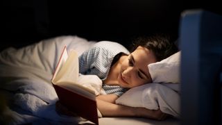 A woman on her side reading a book in bed lit by a lamp