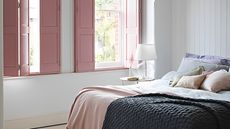 A white bedroom with pink wooden shutters, a pink throw and a glass nightstand light
