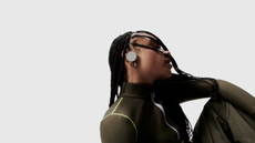 A young person with dreadlocks and a dark green top is wearing an iyO one headphone in their right ear. The rest of the image is a light coloured background