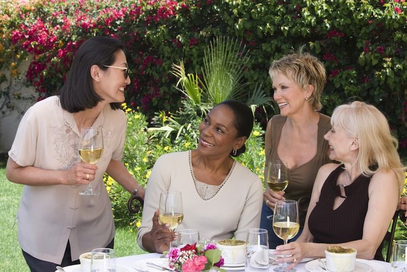 Middle-age women gather around a table, laughing and drinking wine