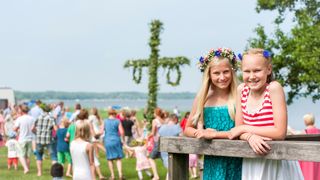 Two girls with flowers in their hair smile in the foreground while people gather around a maypole in the background.