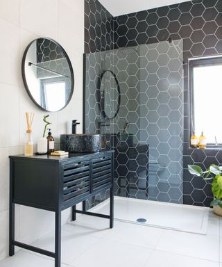 Black hexagon tiled shower enclosure in bathroom with black vanity unit and black round wall mirror